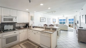Una cocina moderna con armarios blancos, encimeras de granito y una isla grande. Se ve el océano a través de las ventanas.
