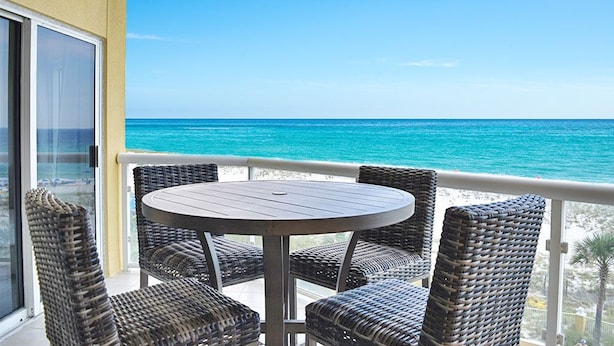 Un balcón con sillas de mimbre y una mesa redonda, con vista al mar turquesa.