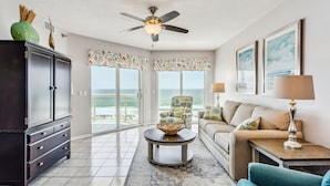 A spacious living room with a large window offering a view of the ocean, a ceiling fan, a dark wooden cabinet, a round coffee table, and a beige sofa. - Great Rates at Emerald Isle 506 - 2B Gulf Front Views! (Pensacola Beach)