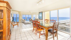 A spacious room with a large dining table, multiple chairs, a wooden cabinet, and large windows offering a view of the beach and ocean. - 3B Gulf Front Paradise at Emerald Isle - 12th Floor (Pensacola Beach)