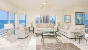 A spacious living room with a large window overlooking a beach, a ceiling fan, and a view of the cityscape. - 3B Gulf Front Paradise at Emerald Isle - 12th Floor (Pensacola Beach)