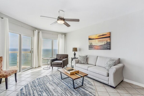 Ein geräumiges Wohnzimmer mit großem Fenster zum Meer, Deckenventilator, Sofa, Couchtisch und einem Gemälde an der Wand.