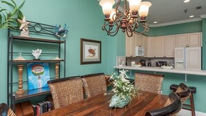 A dining area with a wooden table and wicker chairs, a chandelier, and a kitchen in the background.