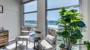 Una habitación moderna con ventanas amplias, un aparador de madera, dos sillones estampados, una mesita auxiliar y una planta verde grande.