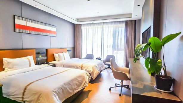Habitación de hotel con dos camas, un escritorio, una silla, un ventanal con cortinas y una planta en maceta.