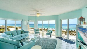 A spacious living room with a large TV, a ceiling fan, and a view of the beach.