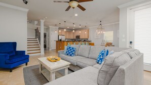 A spacious living room with a grey sofa, blue armchair, and a wooden coffee table. A ceiling fan and pendant lights are visible.
