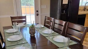 A dining room with a long table set for six, wooden chairs, a black refrigerator, and a window with blinds.