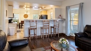 A modern living room with a black leather sofa, a glass coffee table with flowers, and a kitchen area with wooden cabinets and a refrigerator.