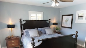 Une chambre à coucher avec un lit en bois foncé, deux tables de chevet avec des lampes, un ventilateur de plafond et une fenêtre avec des volets.