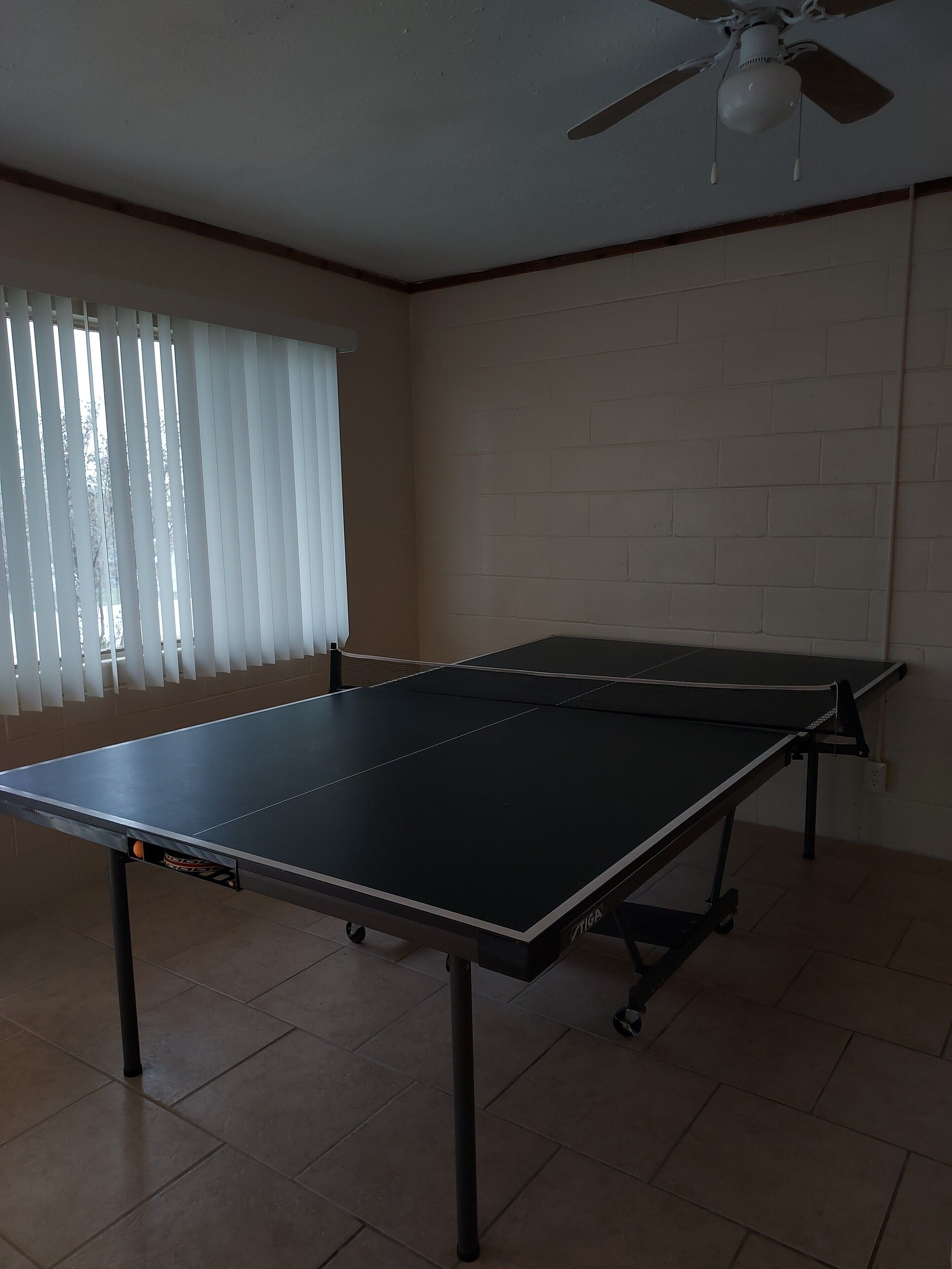 Una stanza con un tavolo da ping pong, una ventola a soffitto e una finestra con tende.