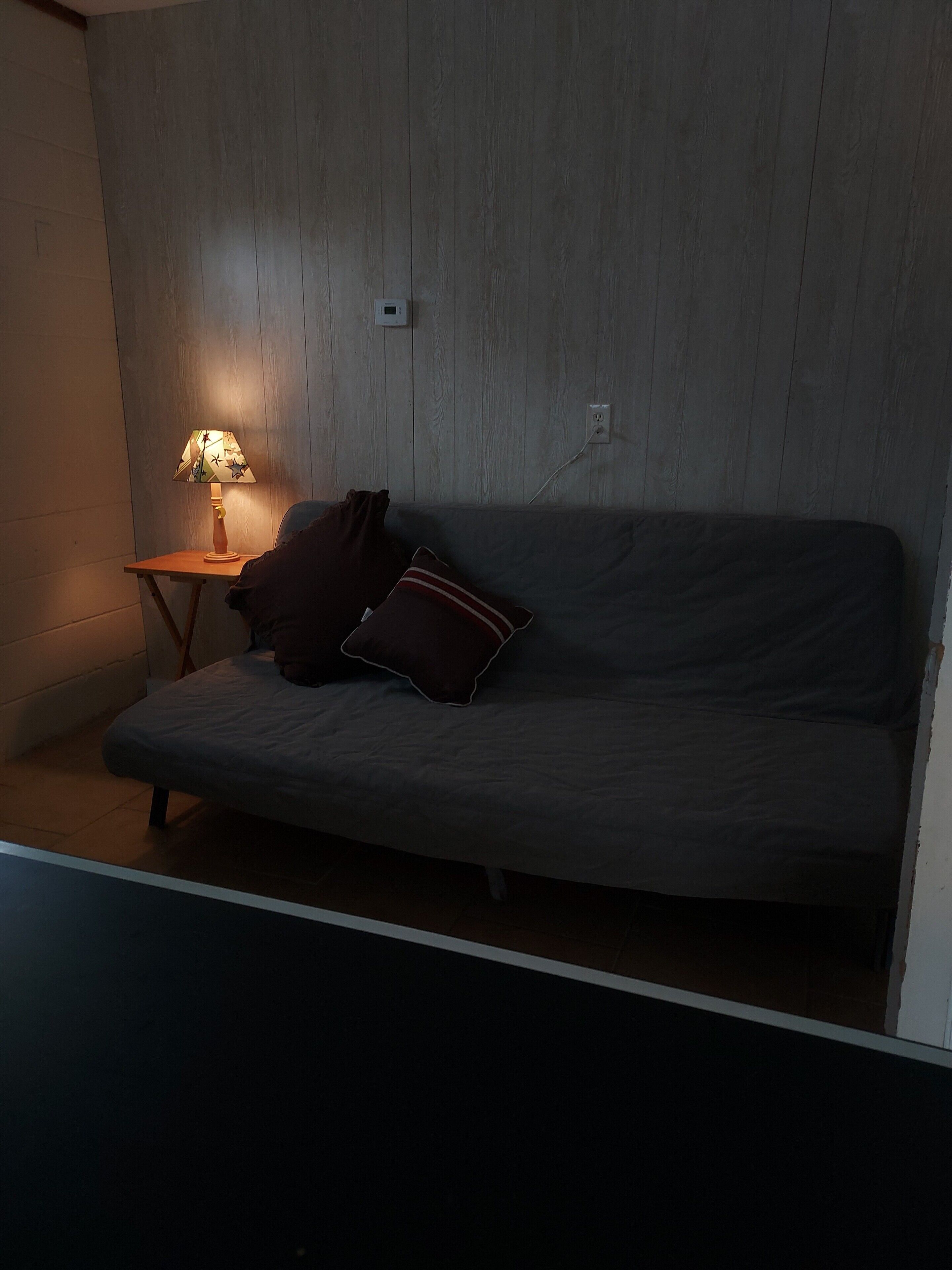 Une pièce avec un canapé gris, une petite table d’appoint en bois sur laquelle se trouve une lampe, et une prise de courant fixée au mur.