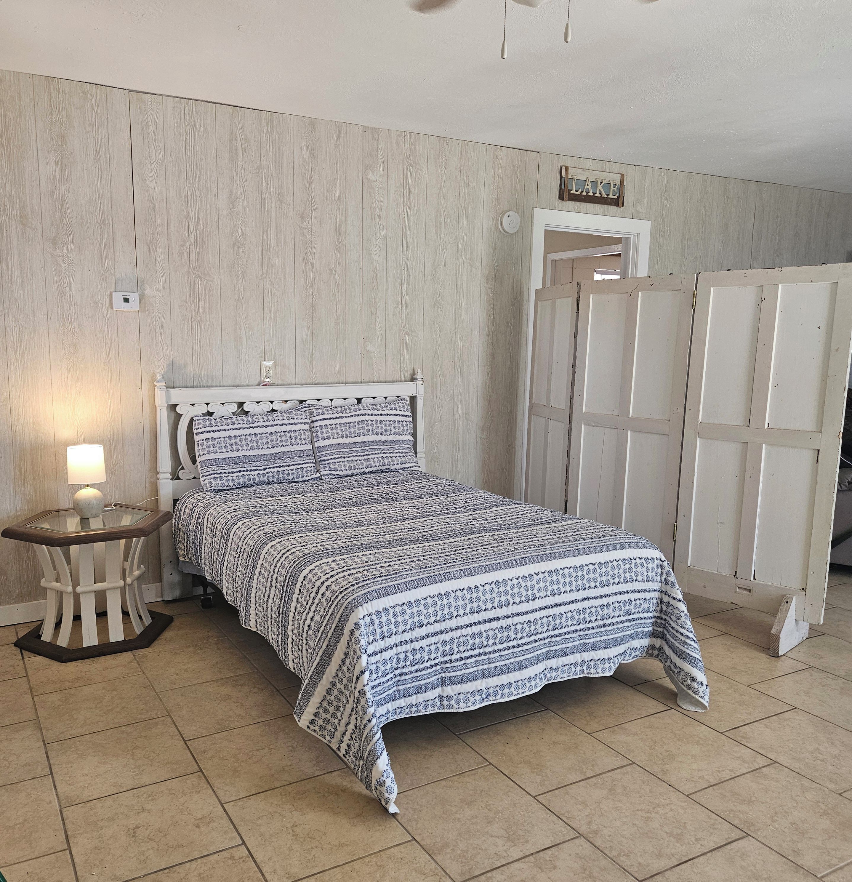 A bedroom with a bed, a bedside table with a lamp, a wardrobe, and a wall-mounted thermostat.