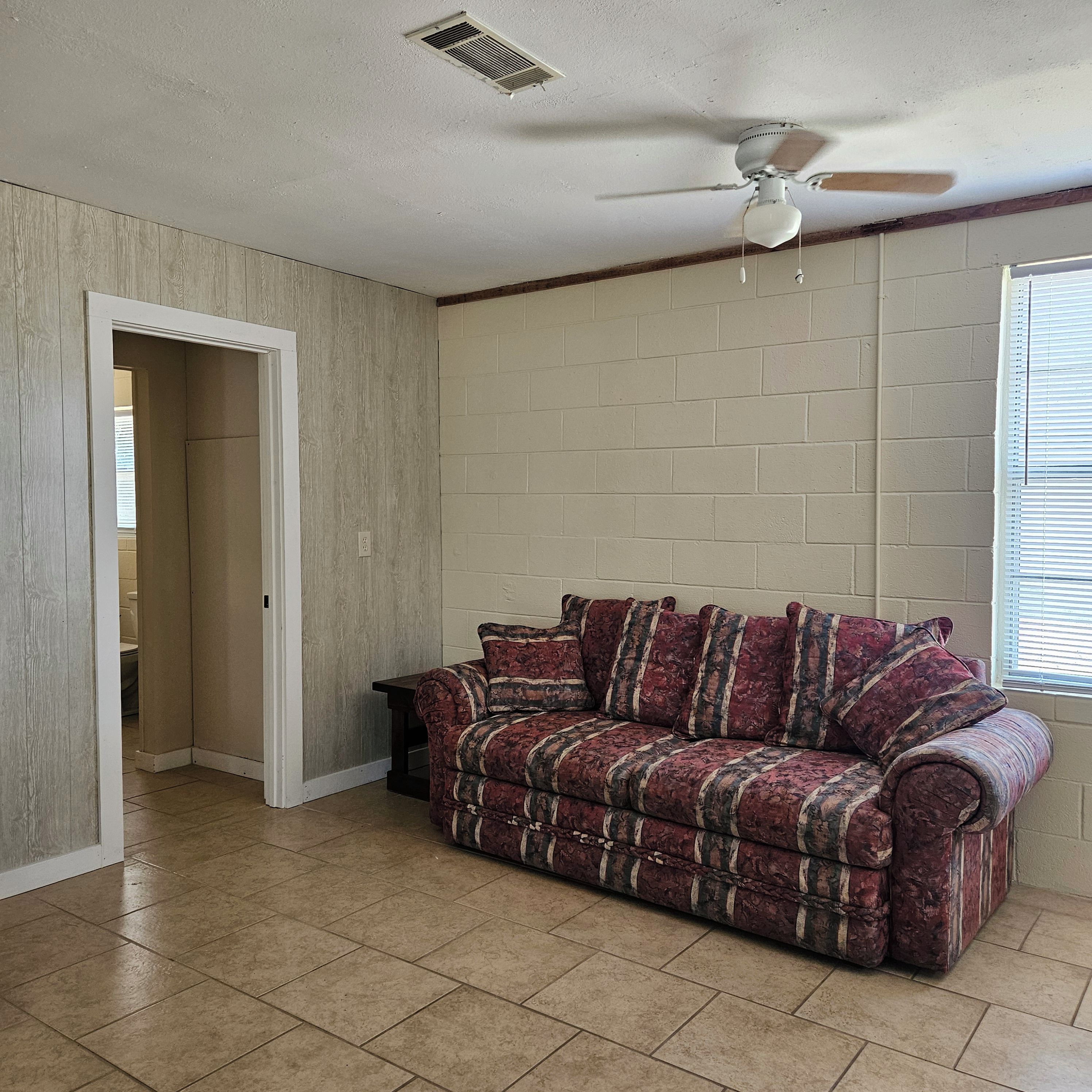A room with a sofa, a ceiling fan, a window with blinds, and a door leading to another room.