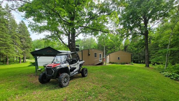 A Polaris RZR ATV parked in a grassy area with a wooden cabin and a metal shed in the background. - Secluded camp off the snowmobile trail (New Sweden)