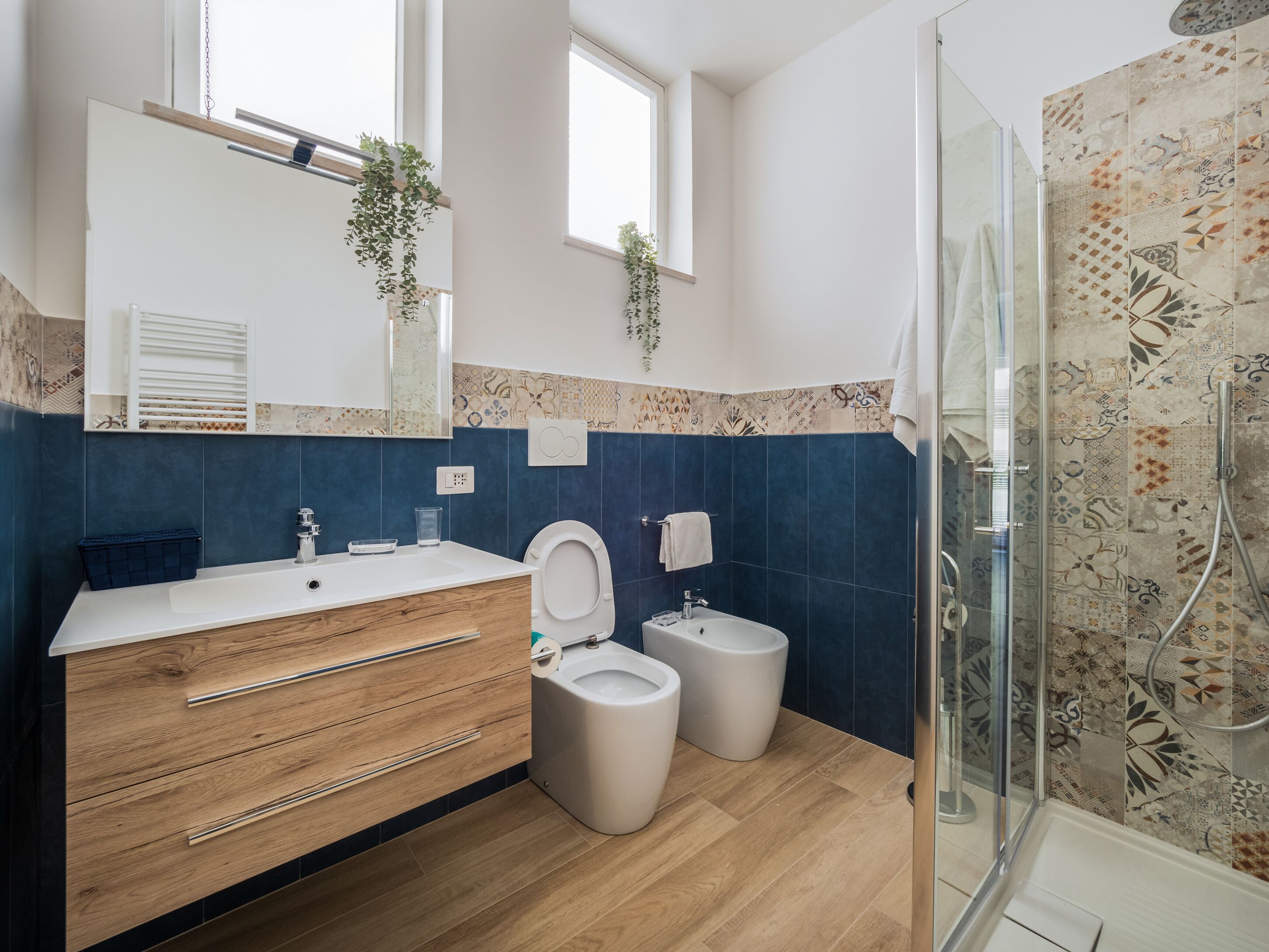 Een moderne badkamer met een houten wastafelmeubel, twee toiletten, een douche en decoratieve tegels.