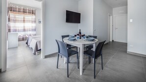 A modern, well-lit room with a dining table set for four, a flat-screen TV mounted on the wall, and a view into another room with a bed and curtains. - Azzurro Park Suite Sorrento (Sorrento)