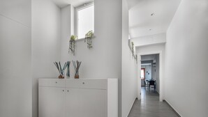 A modern, minimalist hallway with white walls and a grey floor. There are two cabinets with handles, a window with hanging plants, and a door leading to another room. - Azzurro Park Suite Sorrento (Sorrento)