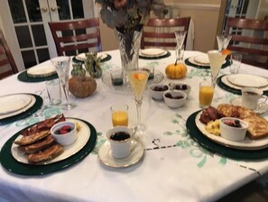 Mesa de café da manhã com bacon, ovos e torradas, além de frutas variadas e bebidas.