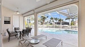 A poolside area with a table and chairs, a ceiling fan, and a view of a swimming pool and palm trees.