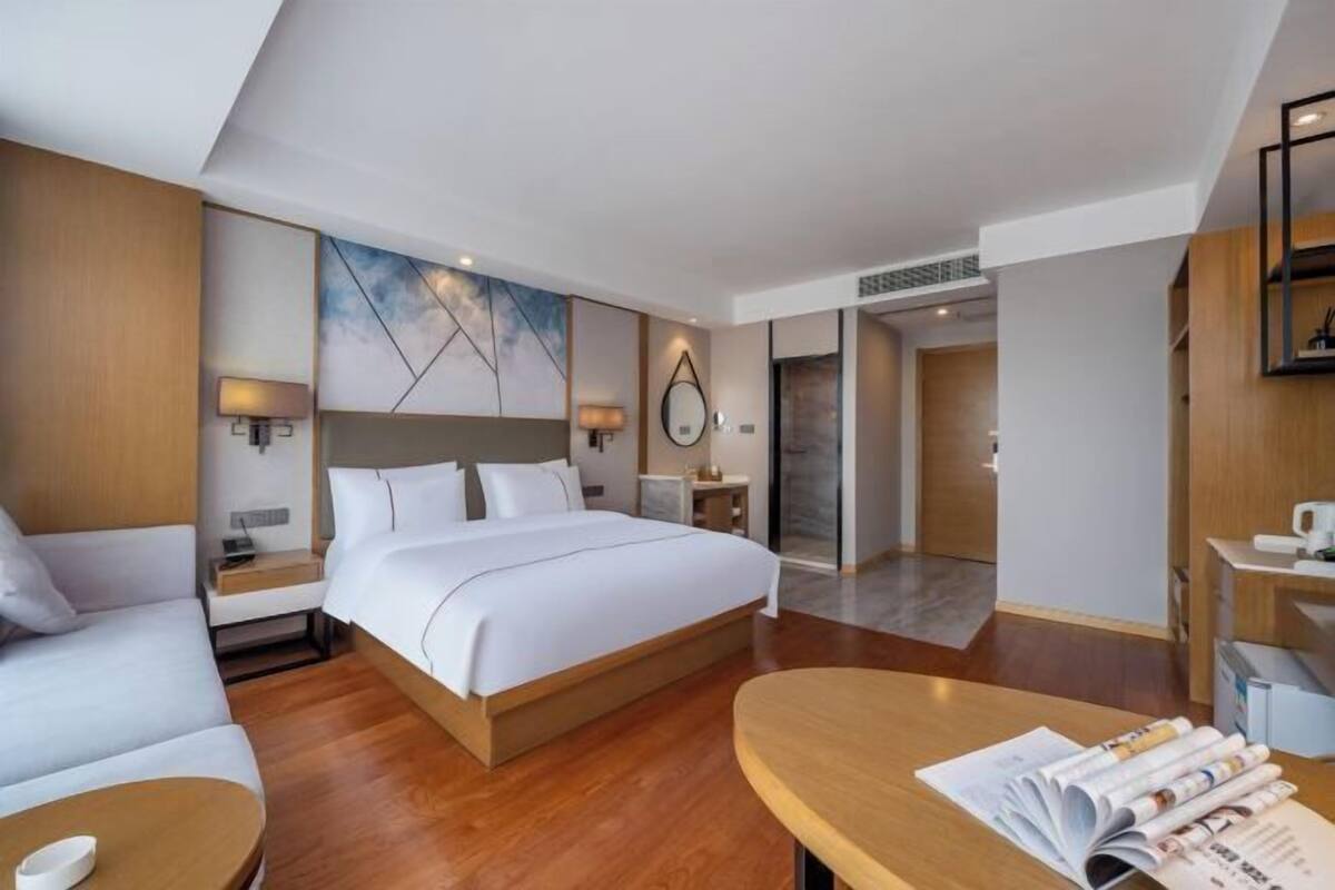 A modern hotel room with a large bed, a desk, a sofa, and a view of the hallway.