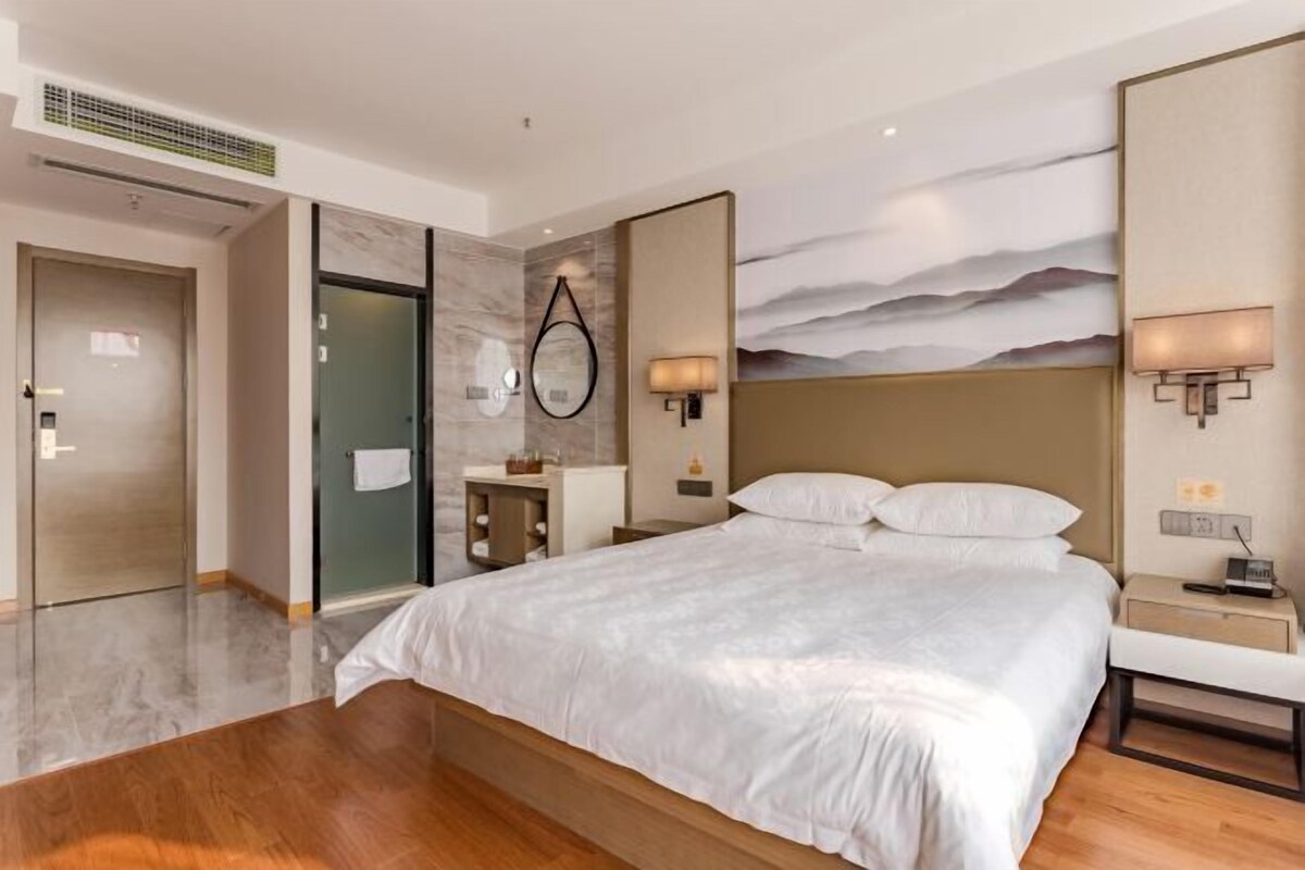 A modern hotel room with a large bed, a bathroom with a glass door, and a wall-mounted artwork.