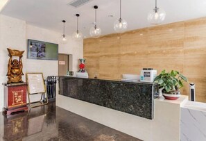 A hotel reception area with a marble counter, hanging pendant lights, a wooden wall, a statue, and a potted plant.