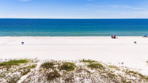 A beach with white sand, a clear blue sea, and sparse vegetation in the foreground. - Luxury Beach House w/ Pool & Stunning Ocean Views (Gulf Shores)
