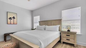 Une chambre à coucher comprenant un lit, des tables de chevet, une commode, une fenêtre avec des stores et un tableau accroché au mur.