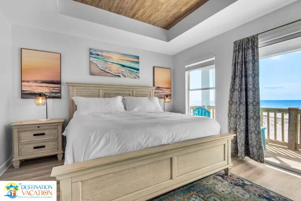 A bedroom with a large bed, two bedside tables, a nightstand, and a view of the beach through a large window.