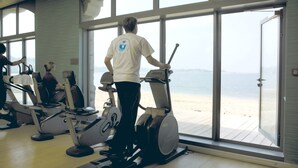 A gym with cardio machines and large windows overlooking a beach.