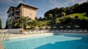 A swimming pool with lounge chairs and umbrellas, a building with balconies, and a landscaped hillside.