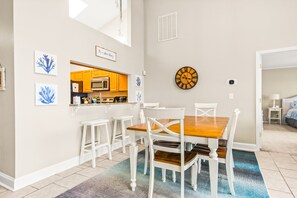 Un comedor con una mesa de madera y sillas blancas, una cocina al fondo y un reloj en la pared.
