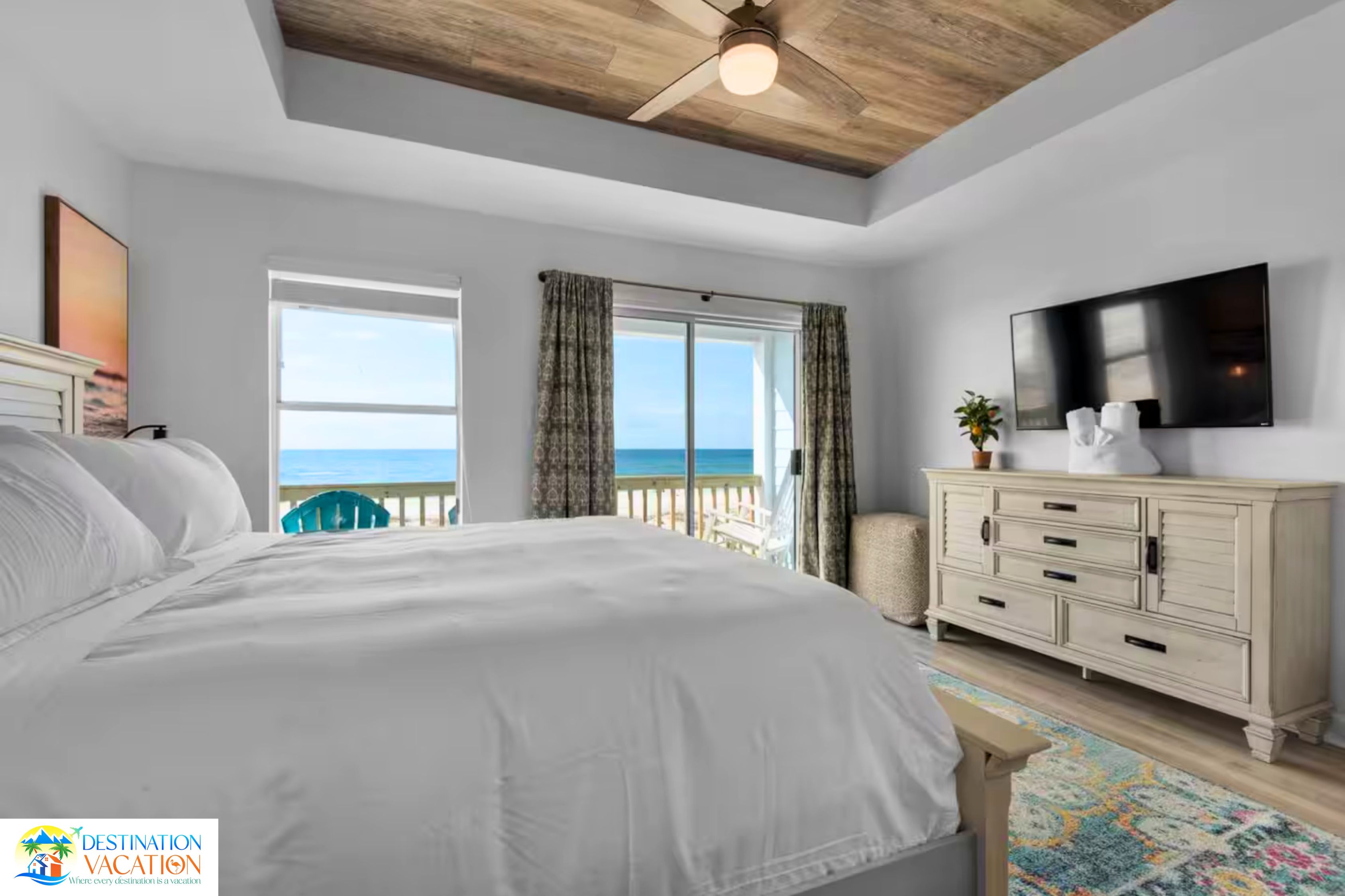 A bedroom with a large bed, a ceiling fan, a TV, and a view of the sea through a sliding door.