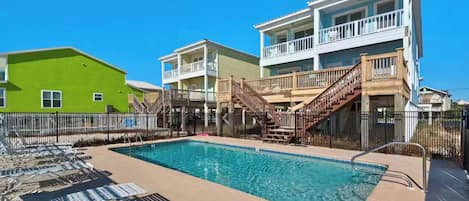 A swimming pool with lounge chairs, a wooden deck, and a multi-story building with balconies.