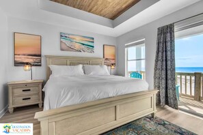 Ein Schlafzimmer mit einem großen Bett, zwei Nachttischen, einem Beistelltisch und Blick auf den Strand durch ein großes Fenster.