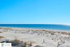 A beach with a clear sky, sandy dunes, and a fenced area with a lounge chair.
