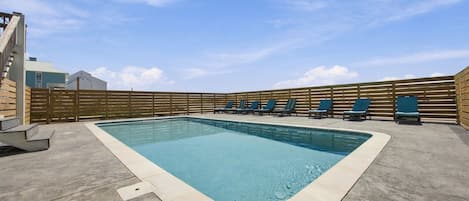 A rectangular swimming pool with a tiled edge, surrounded by a wooden fence and a row of blue lounge chairs.