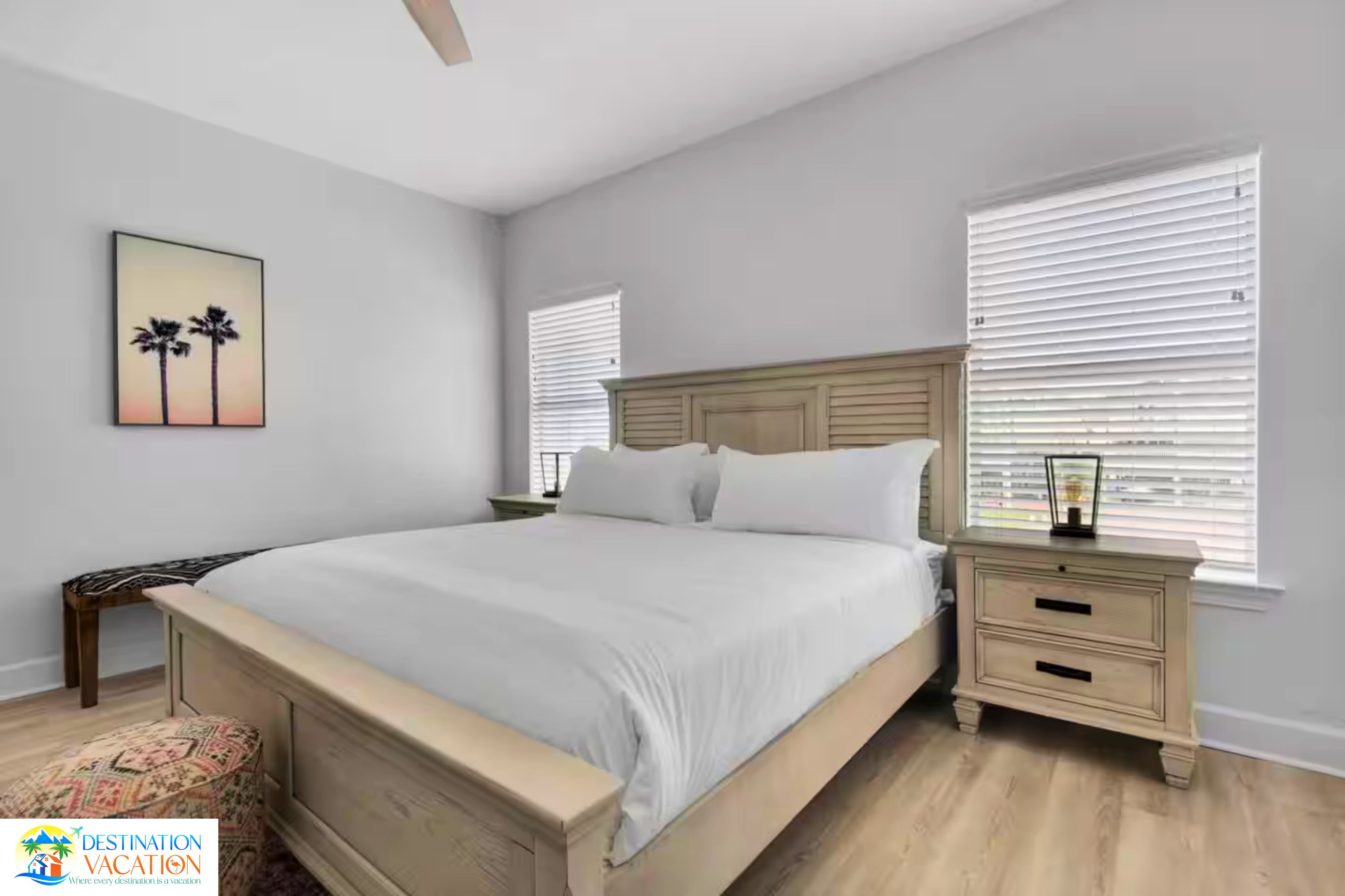 A bedroom with a large bed, two bedside tables, a nightstand, a framed picture on the wall, and a window with blinds.
