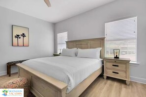 Un dormitorio con una cama grande, dos mesitas de noche, una mesita auxiliar, un cuadro en la pared y una ventana con persianas.