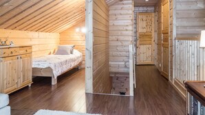 A wooden cabin bedroom with a sloped ceiling, a bed with a patterned bedspread, a wooden dresser, and a small table with a lamp. - Rapsakka a by Interhome (Kittilä)