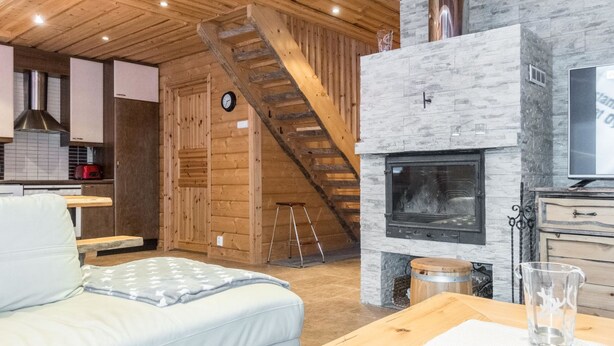 A cozy living room with a sofa, a fireplace, a wooden staircase, and a kitchen area in the background. - Rapsakka a by Interhome (Kittilä)