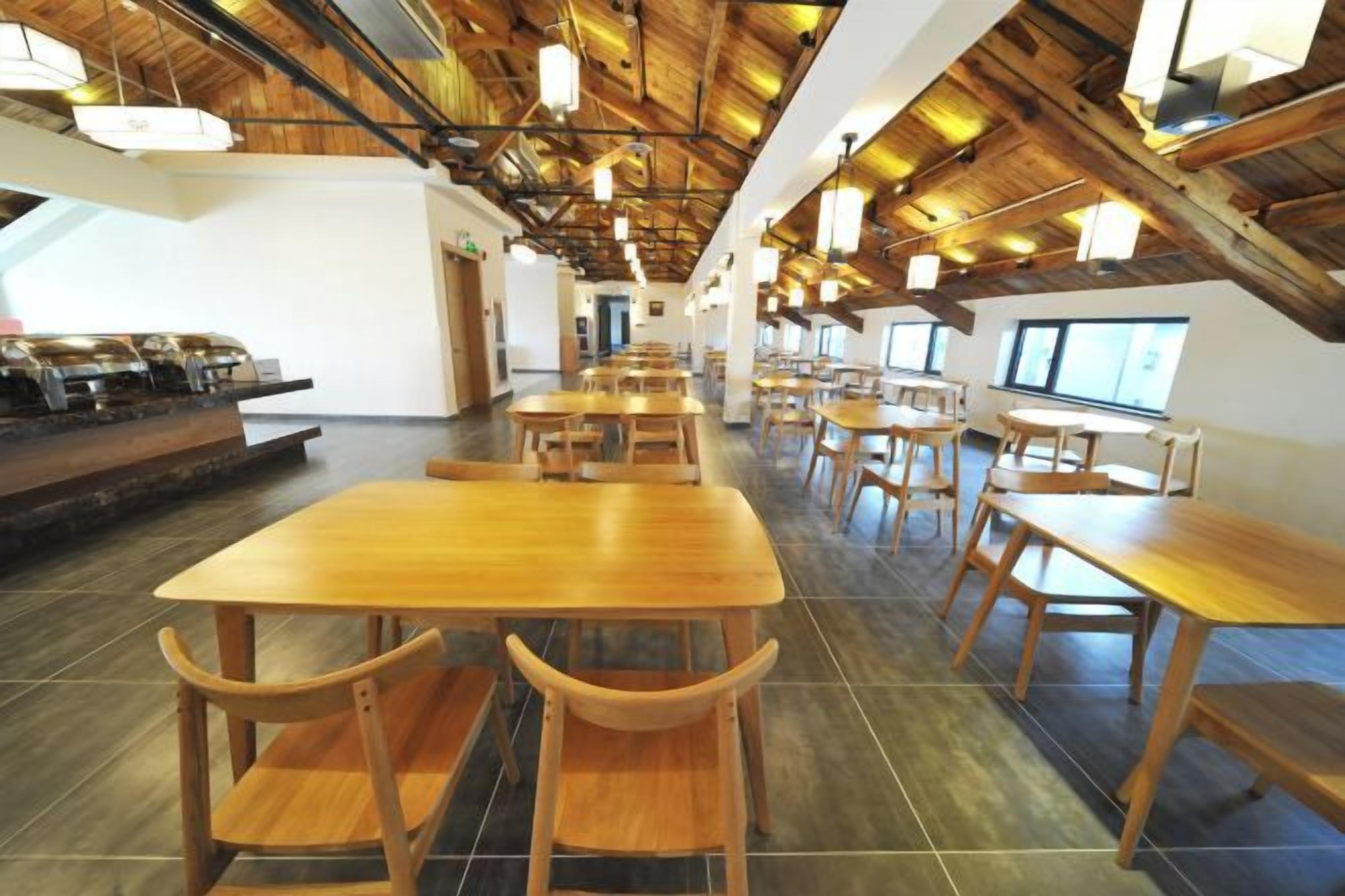 A spacious dining area with long tables and chairs, high ceilings with exposed beams, and large windows allowing natural light.