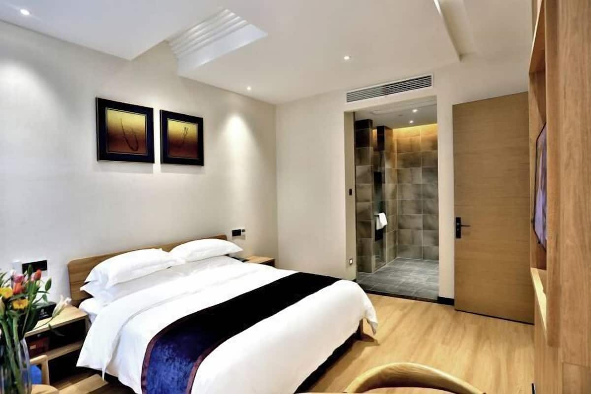 A modern hotel room with a large bed, a bedside table, a nightstand, a shower area, and a wardrobe.