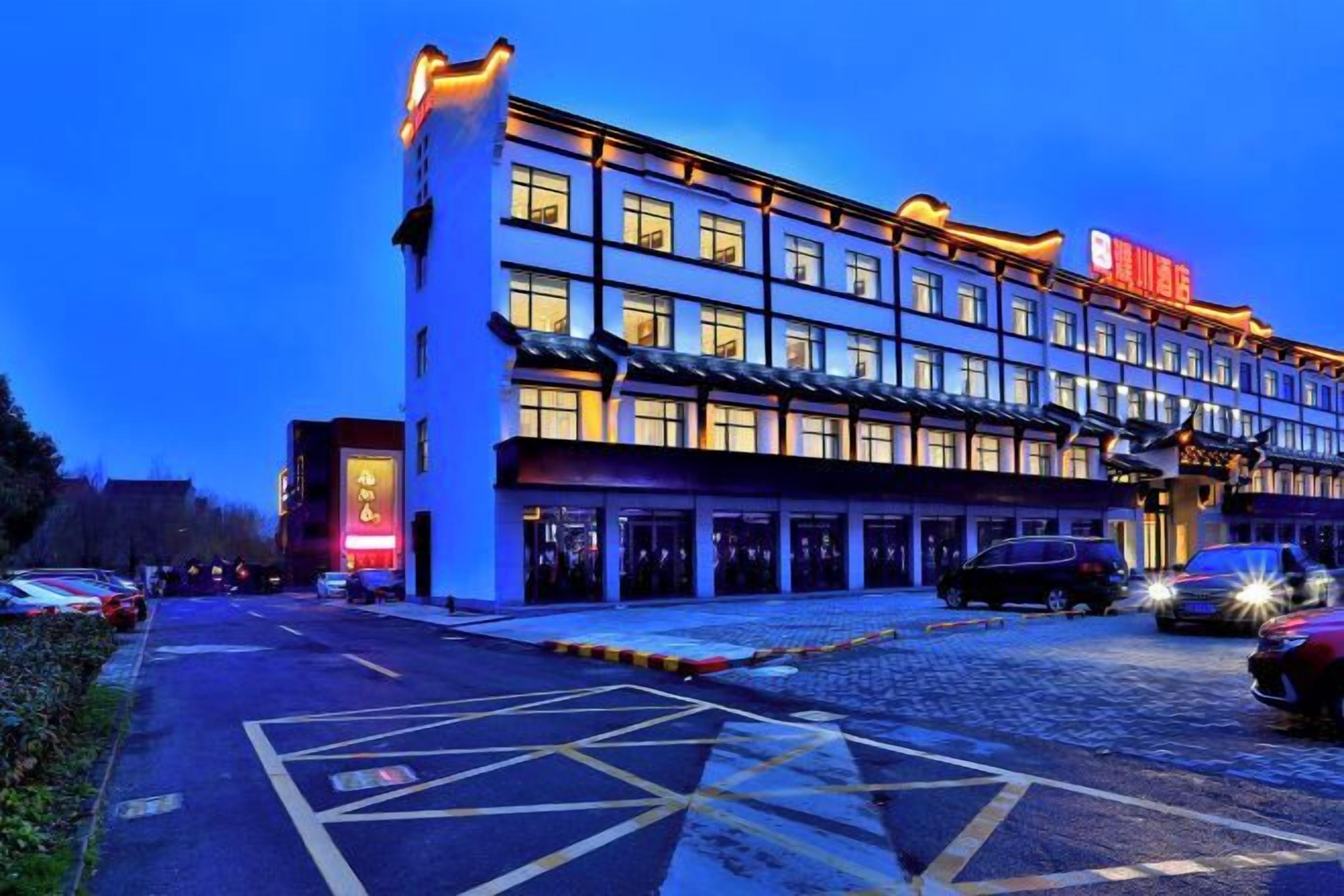 Ein mehrstöckiges Hotel mit einer Fensterreihe im Erdgeschoss, einem Parkplatz mit Markierungen und einem Gebäude mit beleuchteter Beschilderung.