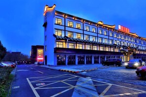 A multi-story hotel with a row of windows on the ground floor, a parking area with marked spaces, and a building with illuminated signage. - Puchuan Hotel (Tongxiang)