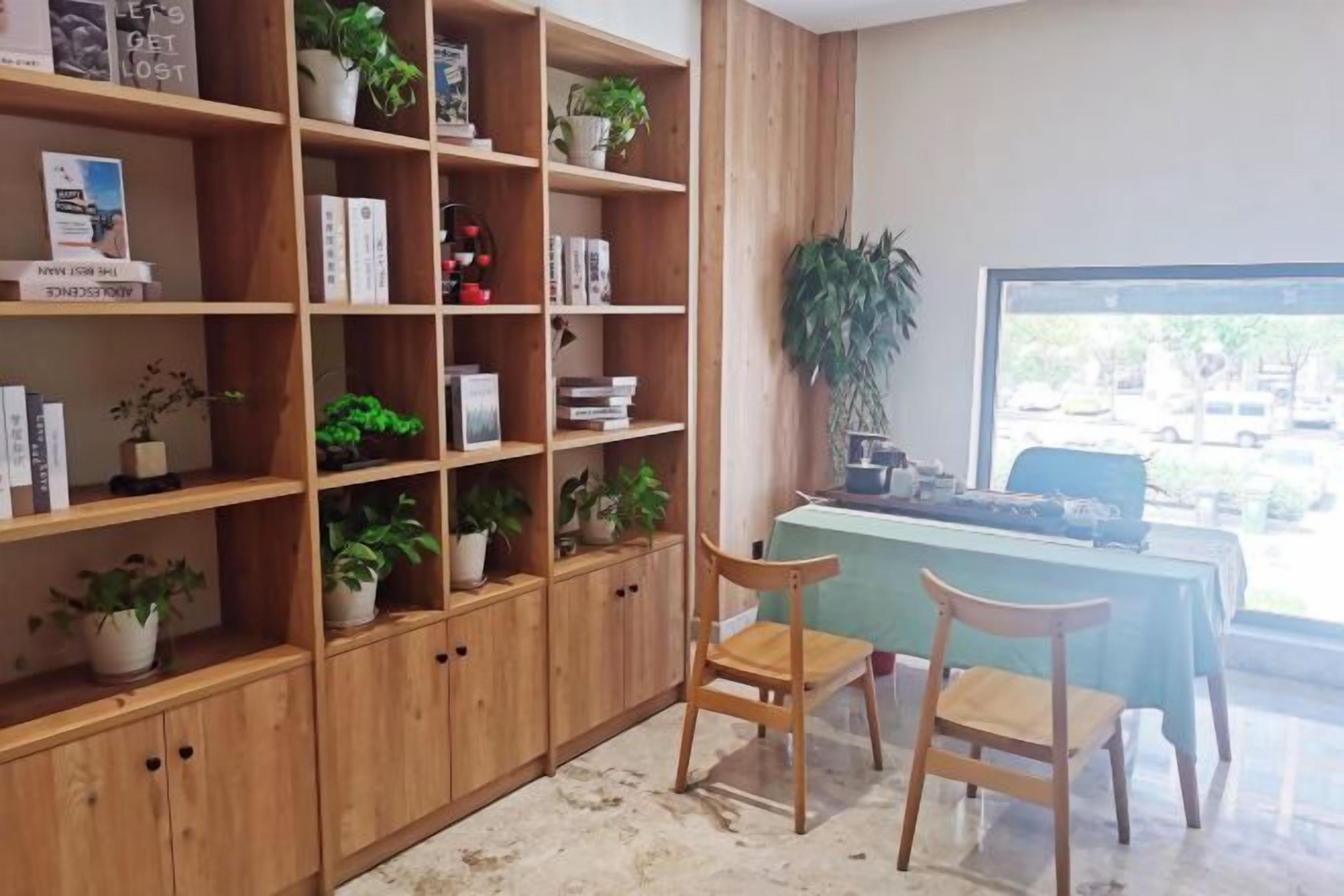 A room with wooden shelving units filled with books and potted plants, a desk with a chair, and a view through a window.