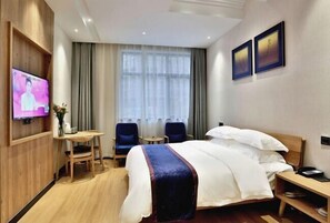 Ein Hotelzimmer mit Bett, Fernseher, Schreibtisch, zwei Stühlen und einem fensterförmigen Öffnung mit Vorhängen.