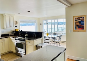 A modern kitchen with white cabinets, a black countertop, and stainless steel appliances. There is a dining area with a glass table and chairs, and a view of a lake through large windows.