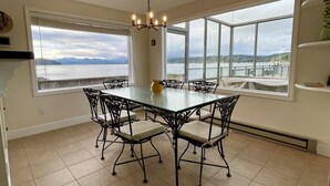 A dining area with a glass table and chairs, large windows with a view of the water, and a chandelier.